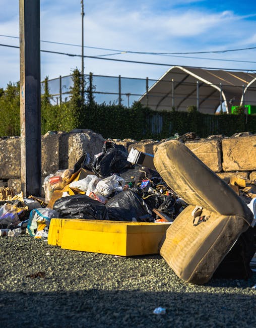 Three large black plastic garbage bags filled with waste are stacked against a dark wooden fence on a street curb. The bags are crumpled and appear to be tightly sealed, with some visible creases and